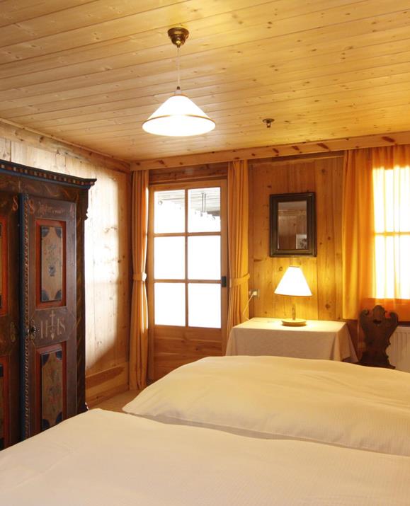 The Church double room: a cozy bedroom with wooden walls and ceiling, a double bed with white linens, warm light from table lamps, and an antique wardrobe adorned with artistic paintings.