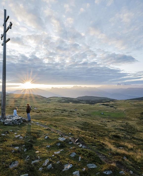 Zwei Personen stehen bei einem hohen Holzkreuz auf einem weiten, grünen Hochplateau und genießen den Sonnenaufgang über den Dolomiten.
