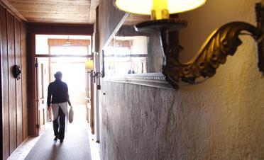Eine Servicekraft geht durch einen gemütlichen, mit Holz verkleideten Flur in Richtung Licht, eine Lampe im Vordergrund spendet warmes Licht.