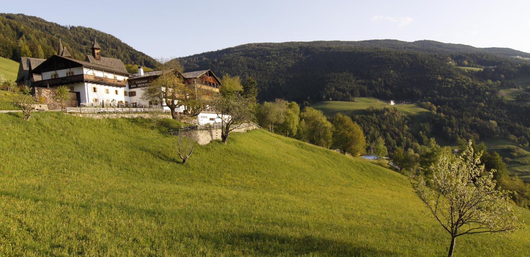 Blick auf den Gasthof Bad Dreikirchen in sonniger Hanglage, umgeben von grünen Wiesen und bewaldeten Hügeln.