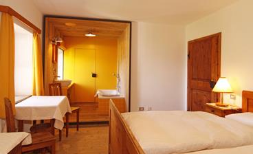 Doppelzimmer Panorama mit integriertem Badbereich mit einer Badewanne und einem Waschbecken, Holzboden und Holzmöbeln, darunter ein Doppelbett, ein kleiner Tisch mit Stühlen sowie Nachttischlampen für eine gemütliche Atmosphäre.