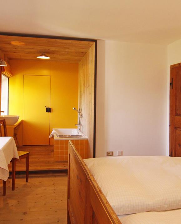 Doppelzimmer Panorama mit integriertem Badbereich mit einer Badewanne und einem Waschbecken, Holzboden und Holzmöbeln, darunter ein Doppelbett, ein kleiner Tisch mit Stühlen sowie Nachttischlampen für eine gemütliche Atmosphäre.