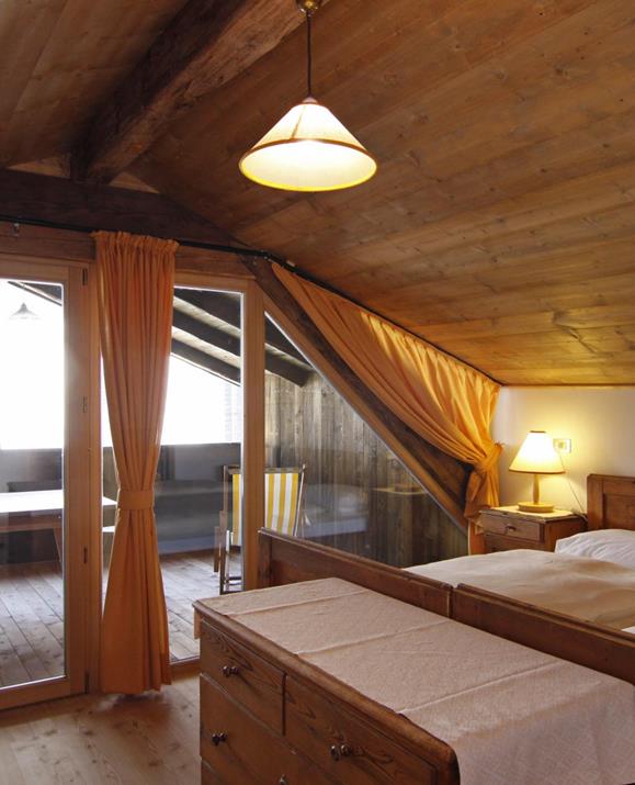 Das Schlafzimmer der Maisonette mit schräger Holzdecke, Holzboden, Zugang zu einem Balkon mit Sitzgelegenheit sowie zwei Einzelbetten mit heller Bettwäsche und gelben Vorhängen vor der Balkontür.