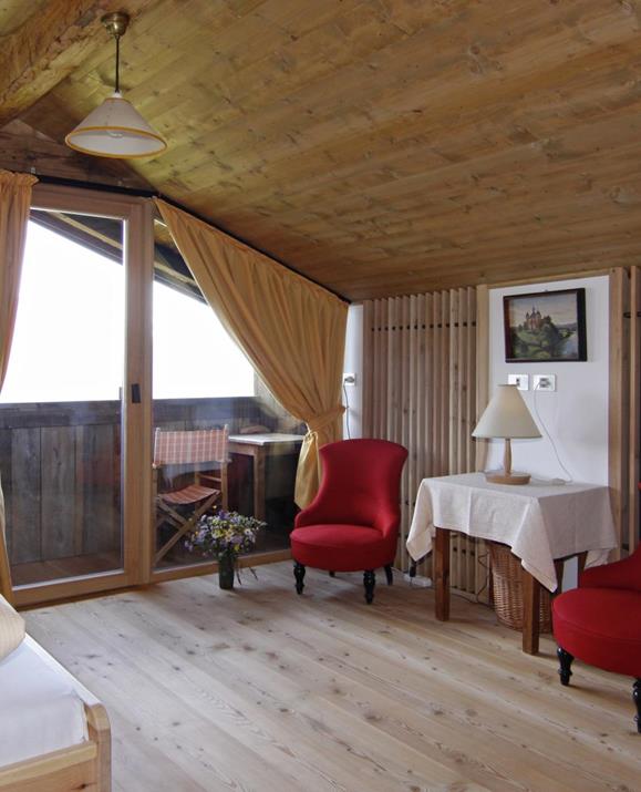 Panorama Superior double room with sloped wooden ceiling, wooden floor, two red upholstered armchairs, a small table with a lamp, access to a balcony with seating, a double bed with light bedding, and yellow curtains in front of the balcony door.