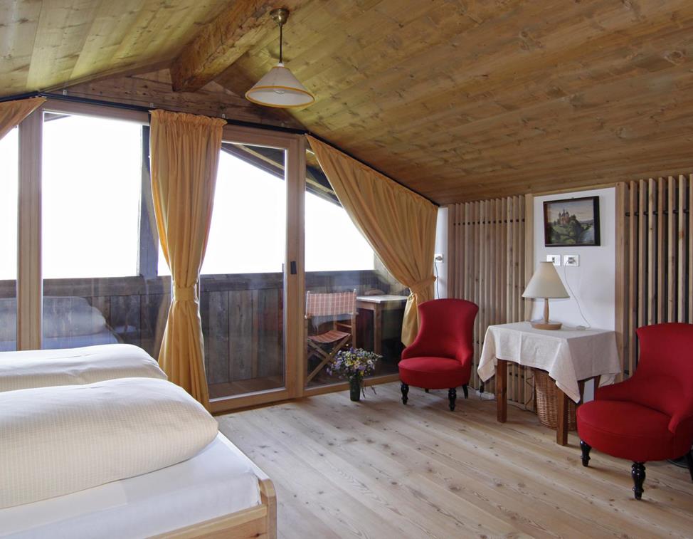Panorama Superior double room with sloped wooden ceiling, wooden floor, two red upholstered armchairs, a small table with a lamp, access to a balcony with seating, a double bed with light bedding, and yellow curtains in front of the balcony door.