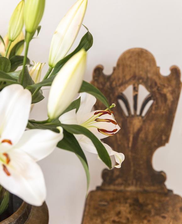 A bouquet of white lilies with closed and open blossoms next to a rustic wooden chair with a carved backrest.