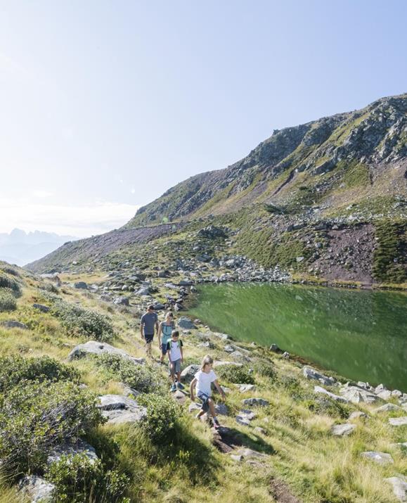 Eine Familie wandert bei Sonnenschein entlang eines klaren Bergsees - dem Totensee - mit grüner Wasseroberfläche, umgeben von Felsen, Gras und sanften Hügeln in alpiner Landschaft.