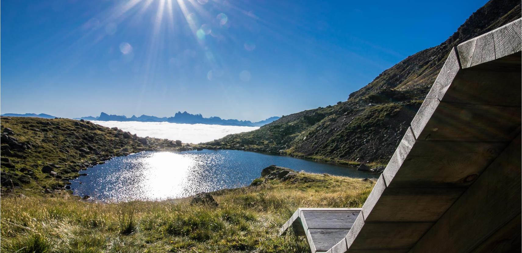 Aussicht auf einen Bergsee mit glitzernder Wasseroberfläche, umgeben von grüner Almwiese und Bergen, im Vordergrund eine hölzerne Relaxliege unter der strahlenden Morgensonne.