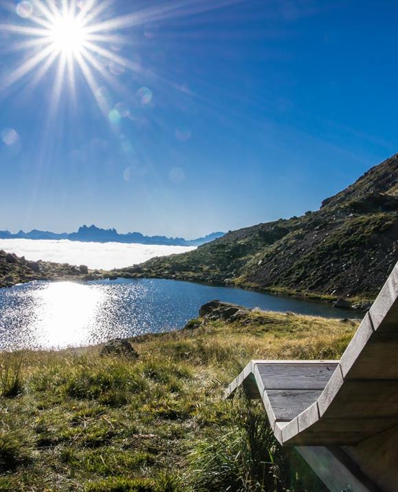 View of a mountain lake with a sparkling water surface, surrounded by green alpine meadows and mountains, with a wooden lounger in the foreground under the bright morning sun.