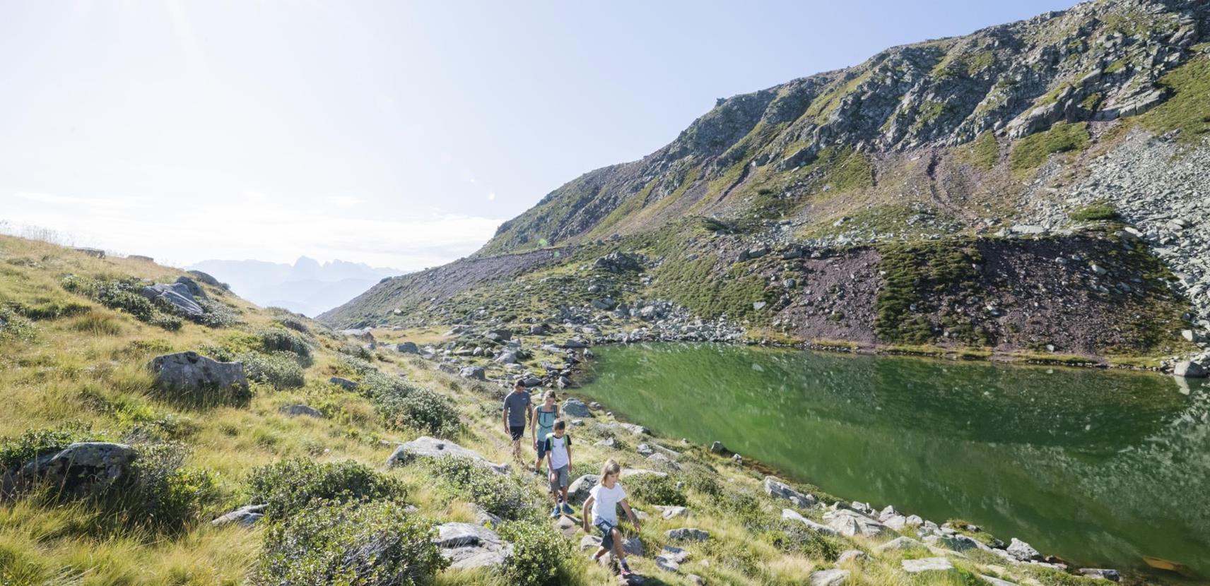 Eine Familie wandert bei Sonnenschein entlang eines klaren Bergsees - dem Totensee - mit grüner Wasseroberfläche, umgeben von Felsen, Gras und sanften Hügeln in alpiner Landschaft.