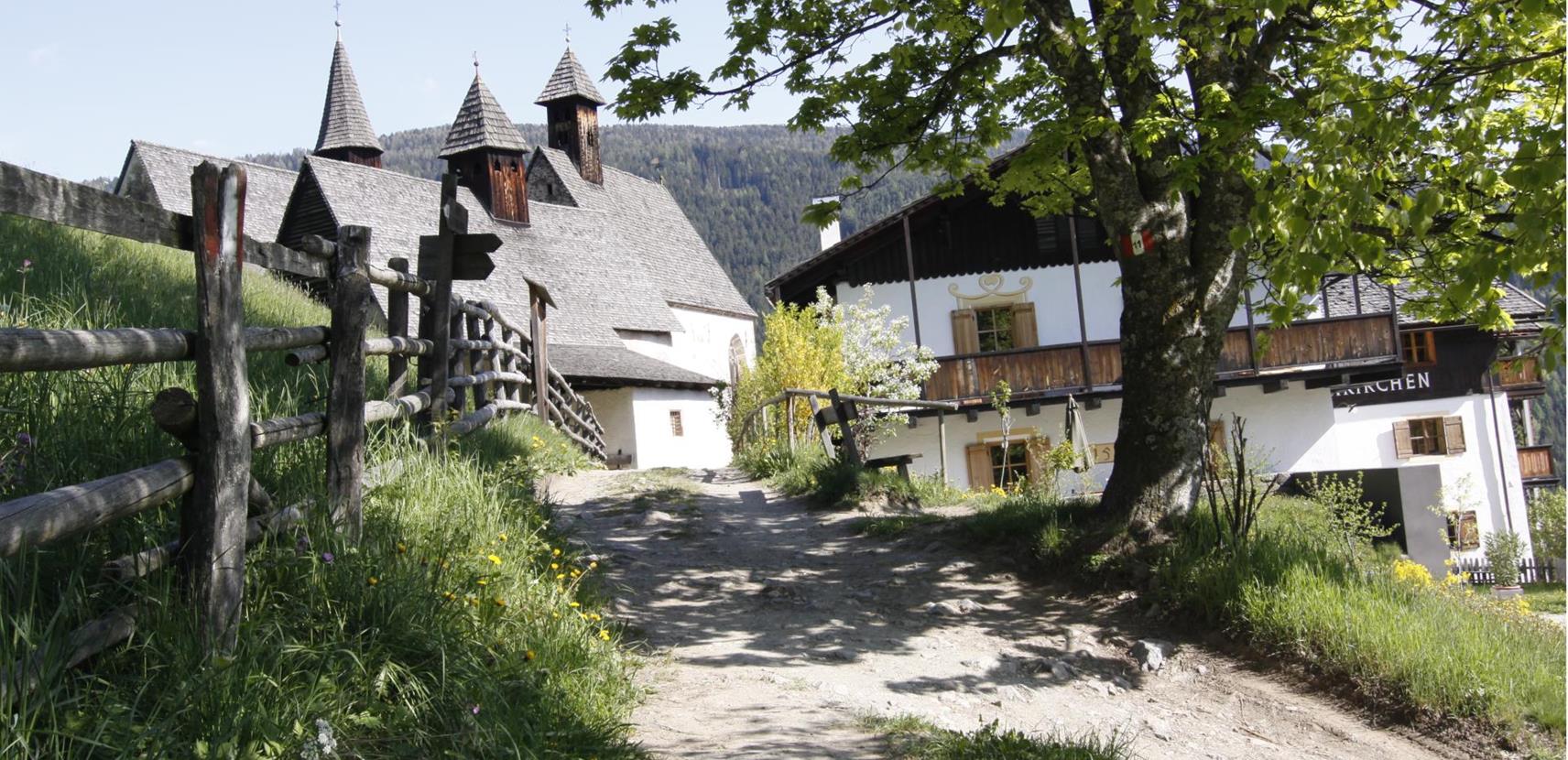 Blick auf die drei Kirchen von Bad Dreikirchen mit unserem traditionellem Gasthof im Frühling, flankiert von einem Holzzaun, grünem Gras und einem blühenden Baum.