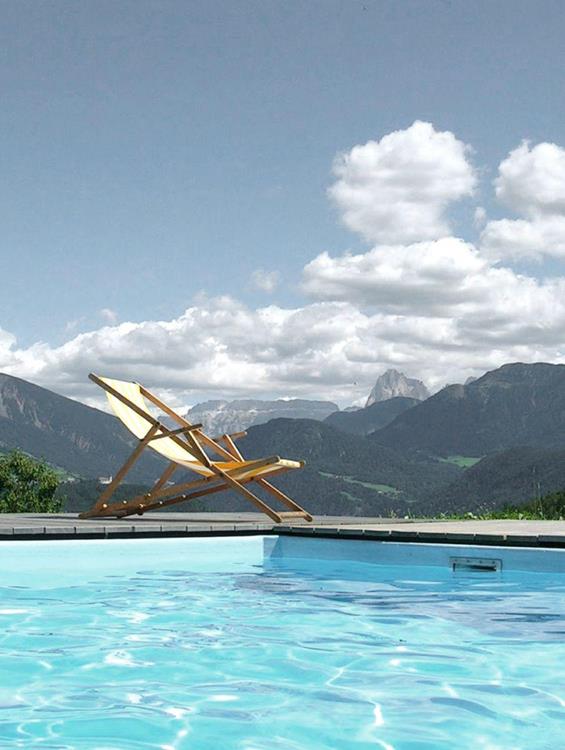 Unser Freischwimmbad mit Blick auf die Dolomiten, im Vordergrund eine Sonnenliege unter blauem Himmel mit vereinzelten Wolken.