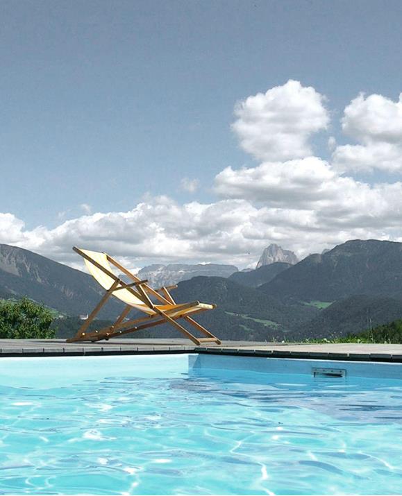 Unser Freischwimmbad mit Blick auf die Dolomiten, im Vordergrund eine Sonnenliege unter blauem Himmel mit vereinzelten Wolken.