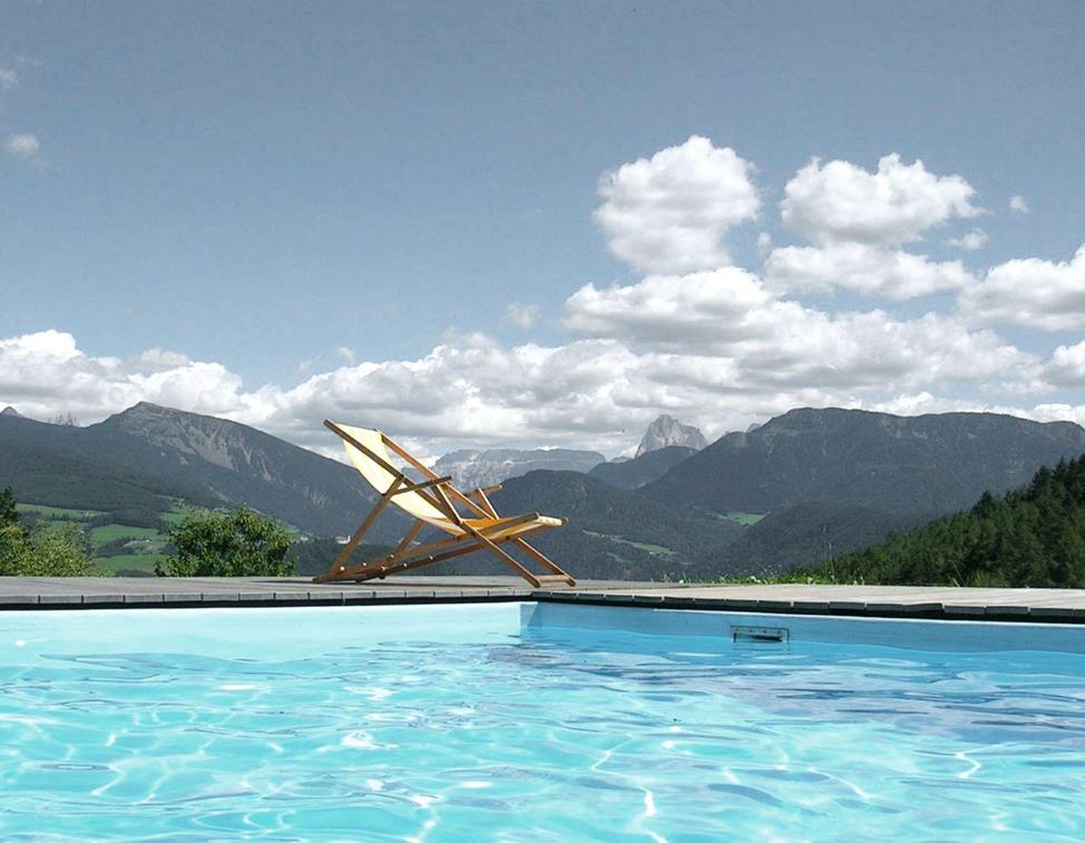 La nostra piscina all'aperto con vista sulle Dolomiti, in primo piano una sdraio sotto un cielo azzurro con qualche nuvola.