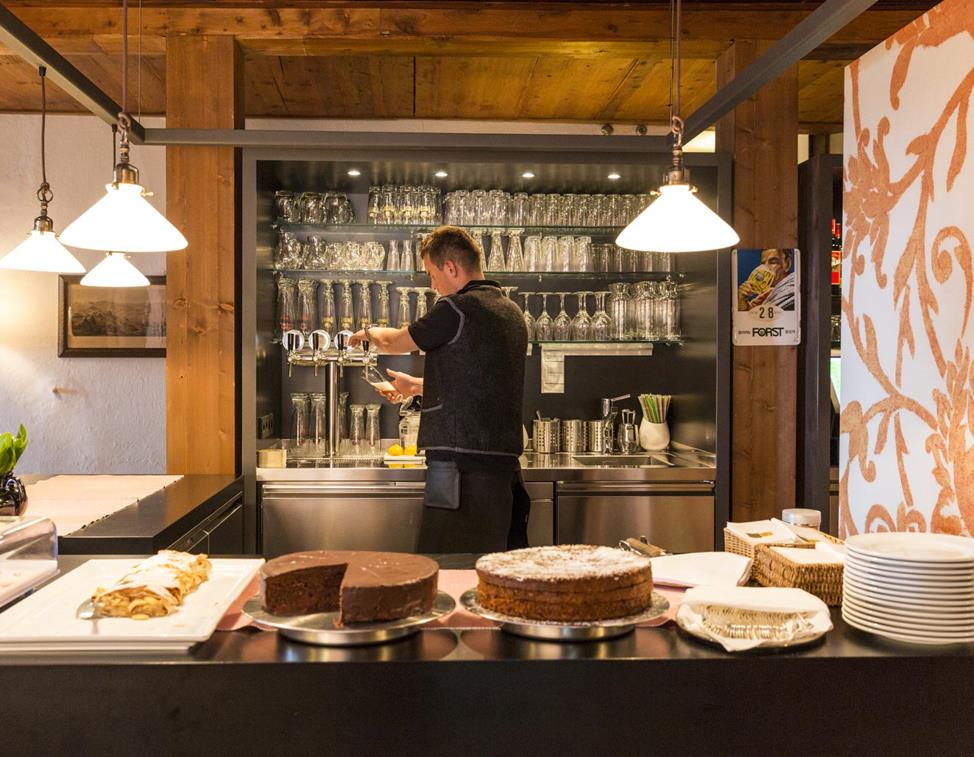 A staff member at the bar preparing a drink; in the foreground a counter with various cakes and desserts