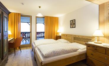 Cozy double room Loggia with wooden floor, rustic wooden bed, bedside tables with lamps, a desk with a red chair, an artistically painted antique wardrobe, and access to a balcony with seating, framed by orange curtains.