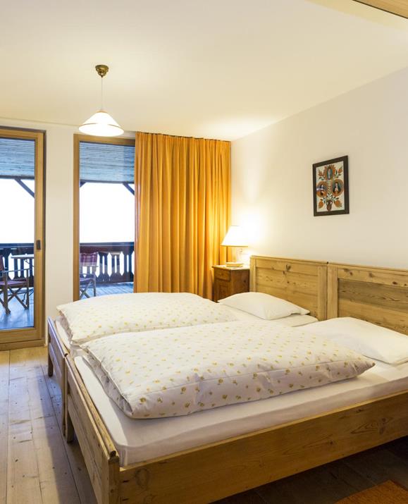 Gemütliches Doppelzimmer Loggia mit Holzboden, rustikalem Holzbett, Nachttischen mit Lampen, einem Schreibtisch mit rotem Stuhl, kunstvoll bemaltem Kleiderschrank und Zugang zu einem Balkon mit Sitzgelegenheit, umrahmt von orangefarbenen Vorhängen.