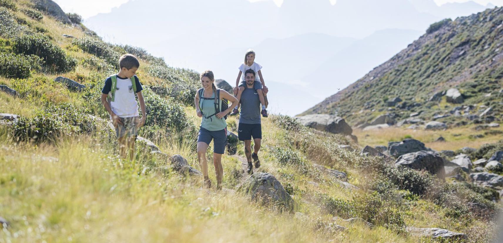 Eine vierköpfige Familie wandert auf einem sonnigen Bergpfad mit Blick auf die Dolomiten im Hintergrund. Ein Mann trägt ein Kind auf den Schultern, während die anderen fröhlich mitwandern.