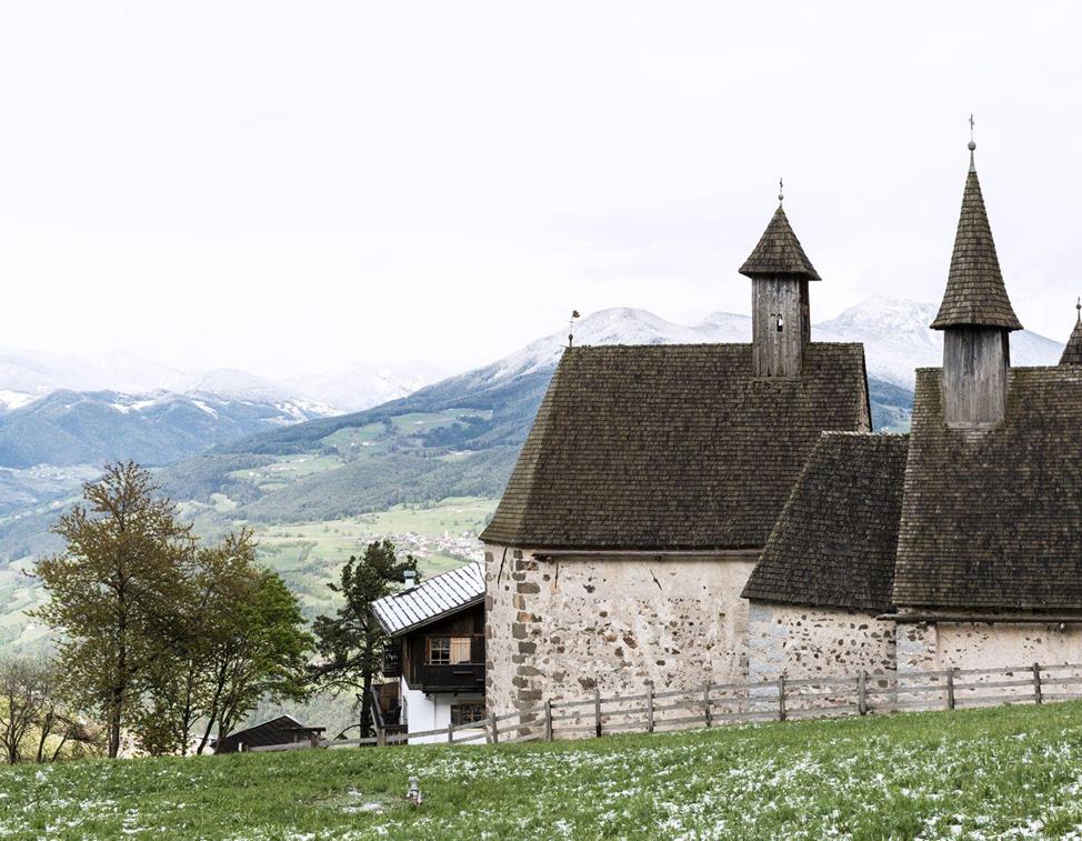 Blick auf die drei Kirchen von Dreikirchen mit ihren charakteristischen Holzschindeldächern vor schneebedeckter Bergkulisse im Frühling.